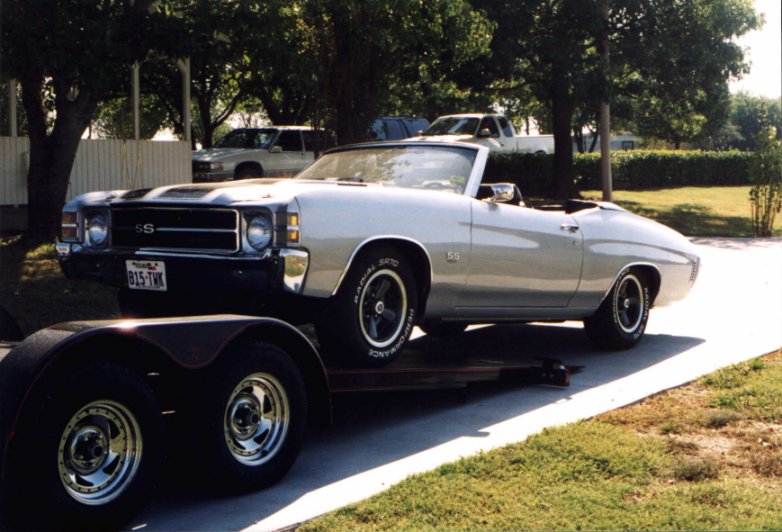 A silver classic Chevrolet convertible car with black stripes is loaded on a trailer in a driveway.