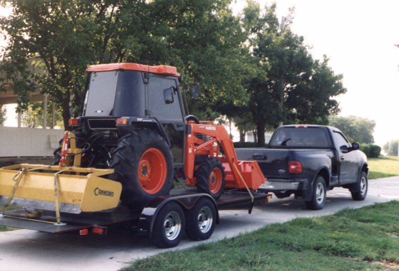 A black pickup truck towing a flatbed trailer carrying an orange tractor and yellow equipment on a driveway.