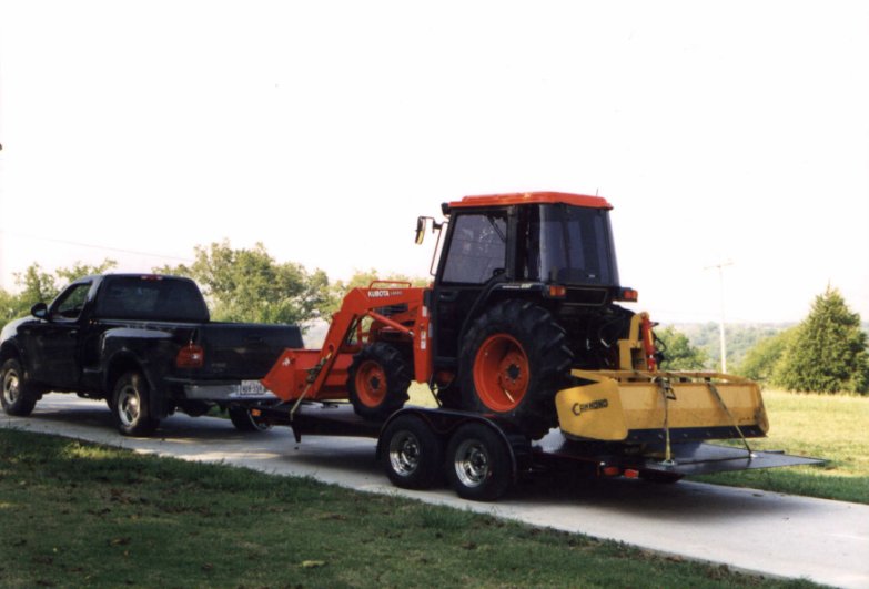 A black pickup truck towing a flatbed trailer carrying an orange tractor with a yellow mower attachment.