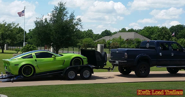 A black pickup truck tows a bright green sports car on a trailer along a suburban driveway.