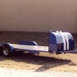 Blue car hauler trailer with dual axles and a white stripe, parked on grass in front of a beige metal building.
