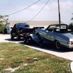 A black truck tows a silver classic convertible car on a trailer up a driveway toward a white garage.