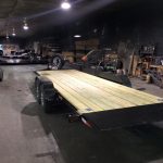 Flatbed utility trailer with wooden deck parked inside a workshop with tools and equipment visible in the background.