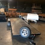A flatbed utility trailer with a spare tire is parked inside a garage with various equipment in the background.