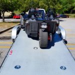 A three-wheeled vehicle loaded onto a flatbed trailer in a parking lot on a sunny day.