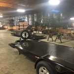 A black metal utility trailer with silver wheels inside a workshop with shelves, tools, and workbenches.
