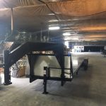 A large gooseneck flatbed trailer is parked inside a spacious warehouse with a high, insulated ceiling.