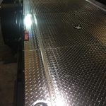 A metal diamond plate trailer bed with two circular latches and a small rectangular object placed on its surface.