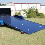 A blue flatbed tilt trailer with dual axles parked on grass near a metal building.