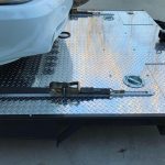 A car's front bumper on a diamond-plate metal flatbed with a securing strap fastened in place.