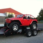 A red Jeep with large off-road tires is secured on a black trailer in a parking lot near a red and white building.
