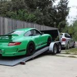 A green sports car is loaded onto a trailer hitched to a silver SUV in a residential driveway.