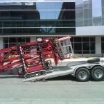 A red scissor lift is being loaded onto a trailer in front of a modern glass building.