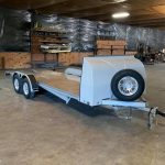 A flatbed trailer with a spare tire is parked inside a large warehouse with shelves and tools in the background.