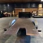 Flatbed car hauler trailer with metal ramp inside a warehouse, surrounded by tires and stacked materials.