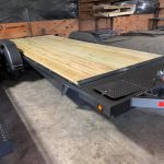 Flatbed utility trailer with a wooden deck in a warehouse with shelves and tools in the background.