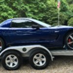 A blue sports car is parked on a trailer in an outdoor setting with trees and grass in the background.