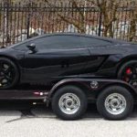 A black sports car is secured on a two-axle trailer parked on the street near a metal fence and trees.